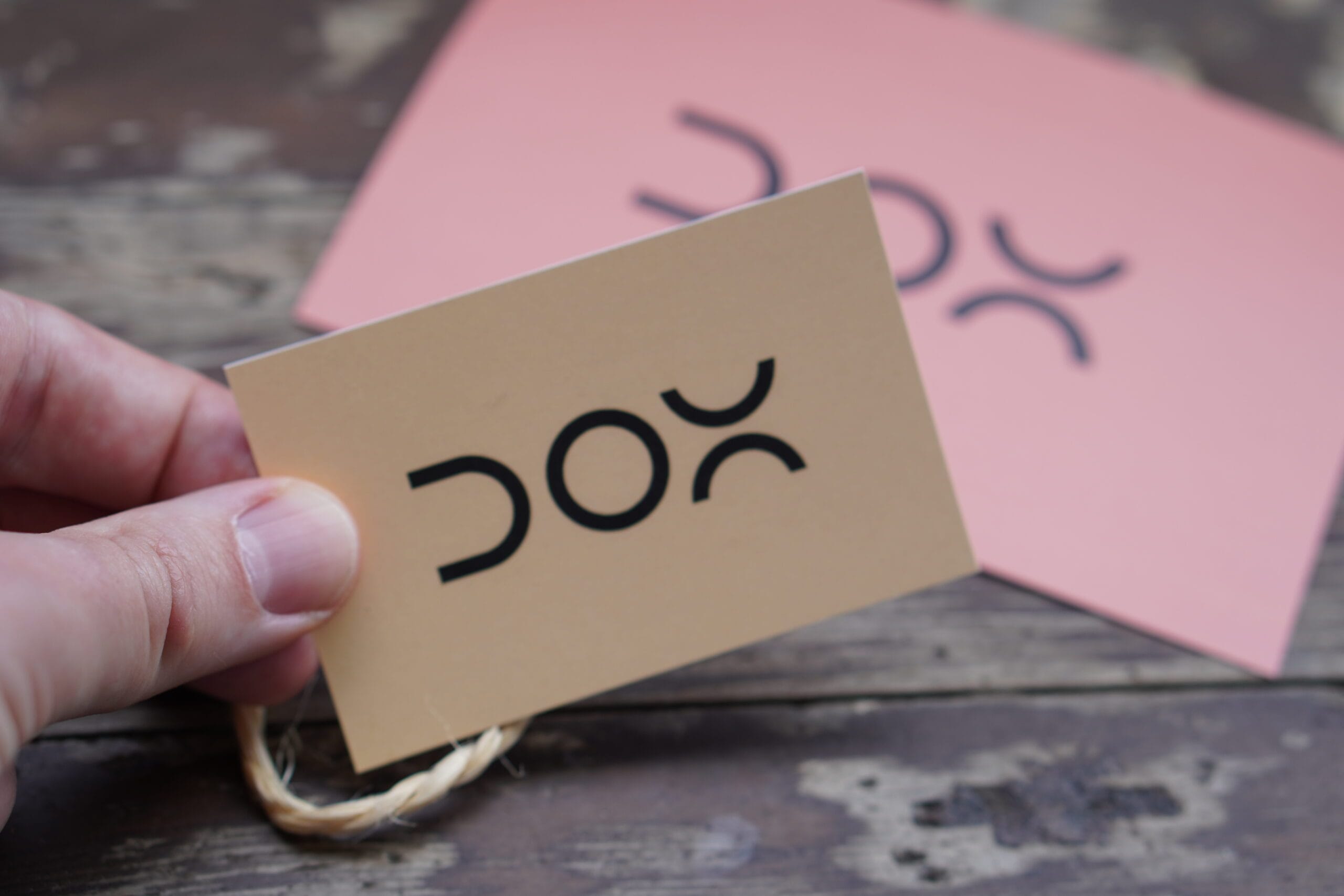 Hand met een bruin visitekaartje van DOX, op de achtergrond wazige roze kaartjes met logo