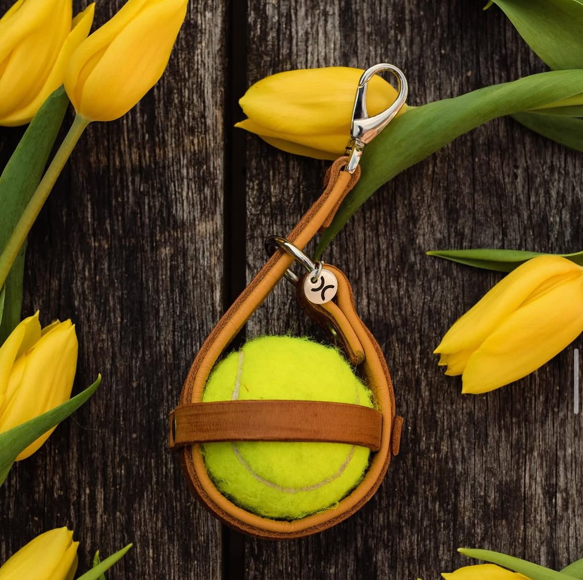 Leren sleutelhanger met een tennisbal erin, tussen gele tulpen op een houten ondergrond