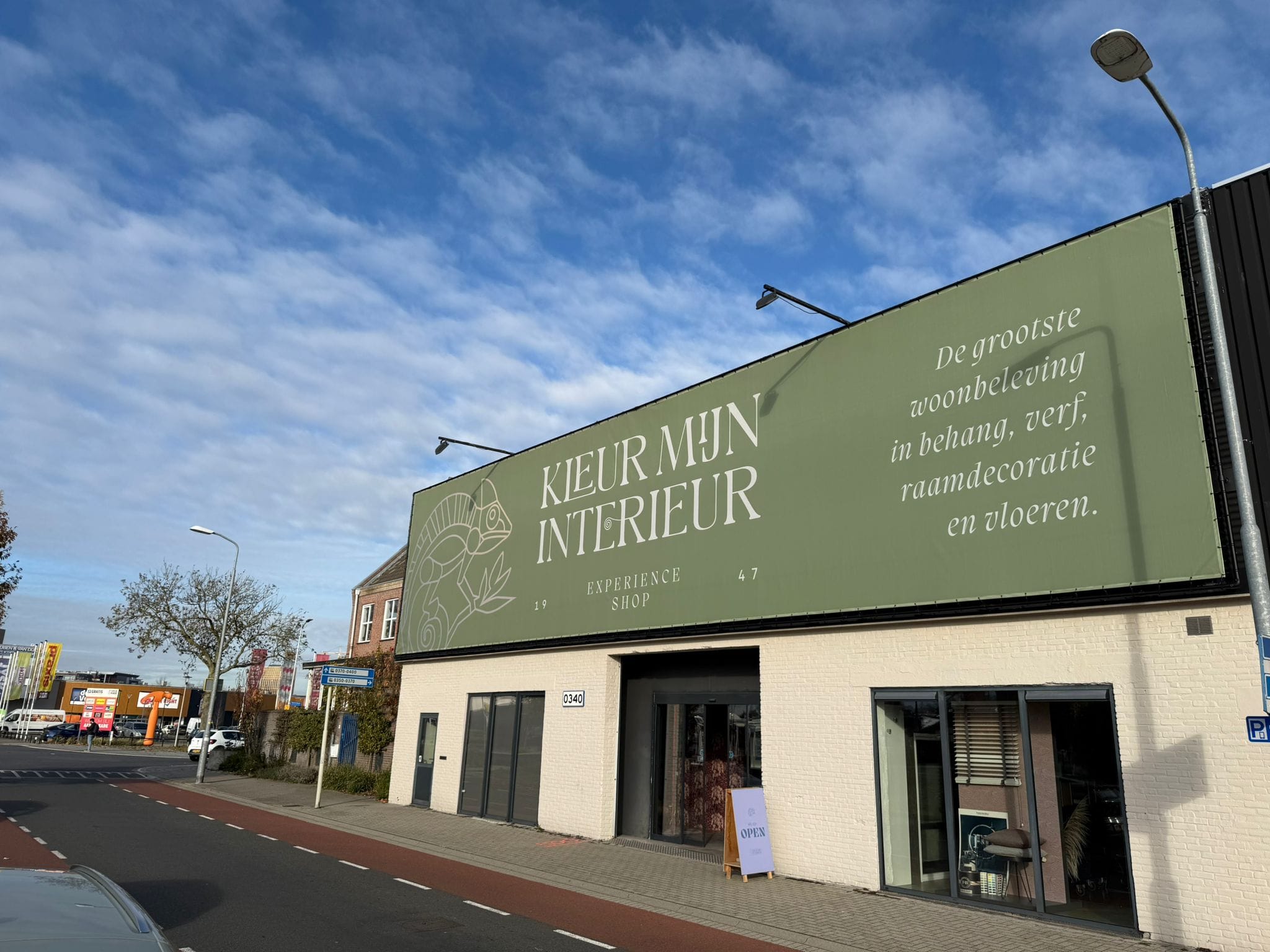 Winkelpand met groene gevel en de tekst Kleur Mijn Interieur Experience shop, onder een blauwe lucht