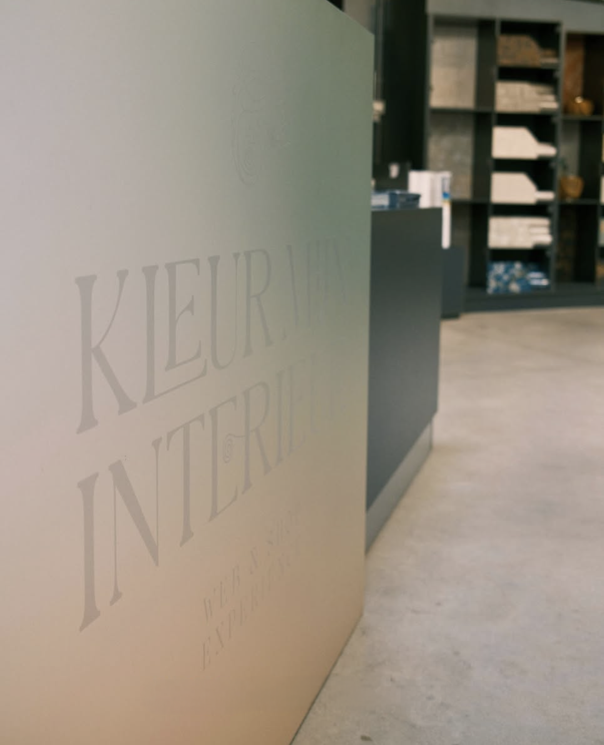 Matglazen balie met gegraveerde naam Kleur Mijn Interieur, schappen met stofstalen op de achtergrond
