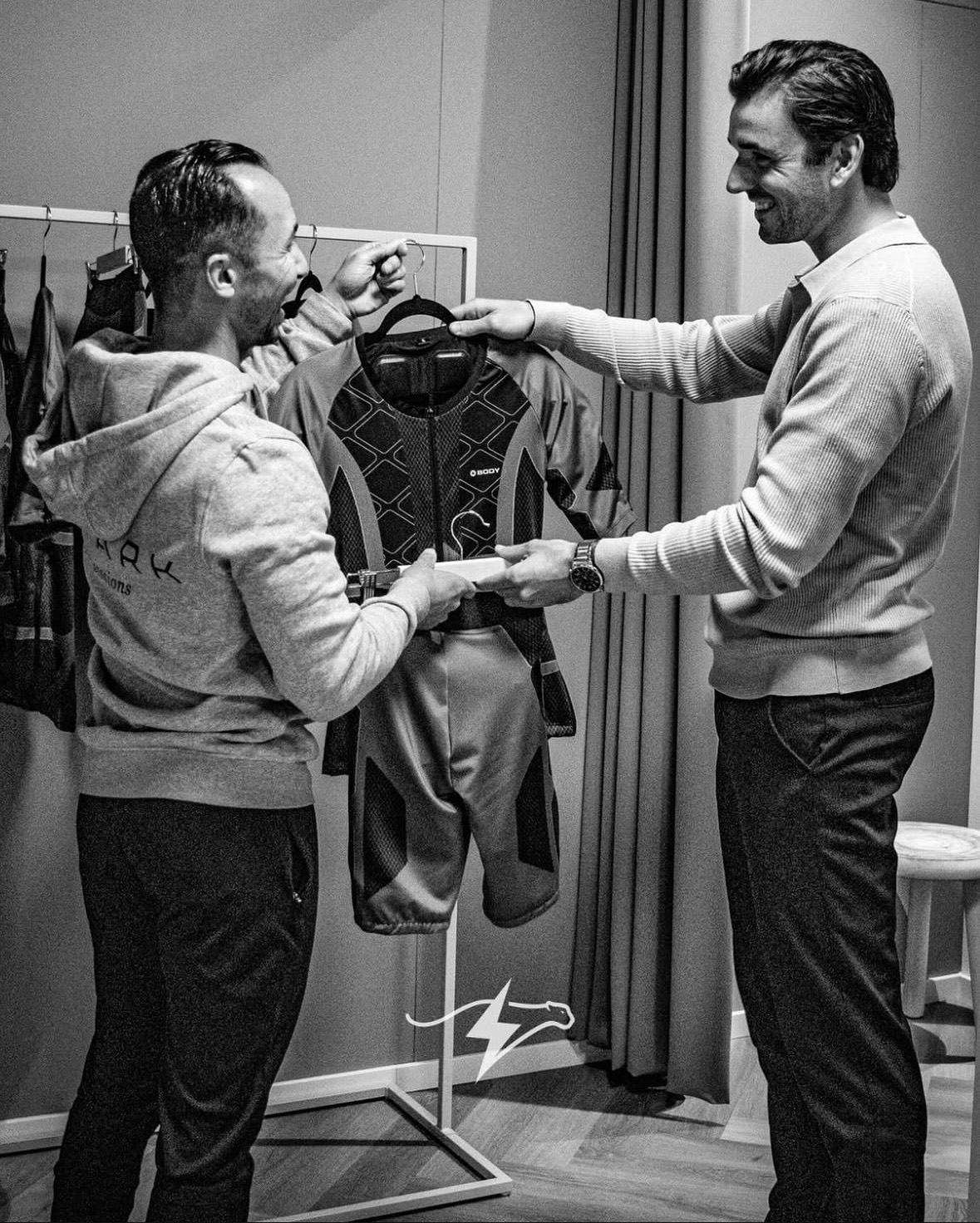 Twee mannen bekijken een sportjack in een paskamer met kledingrek