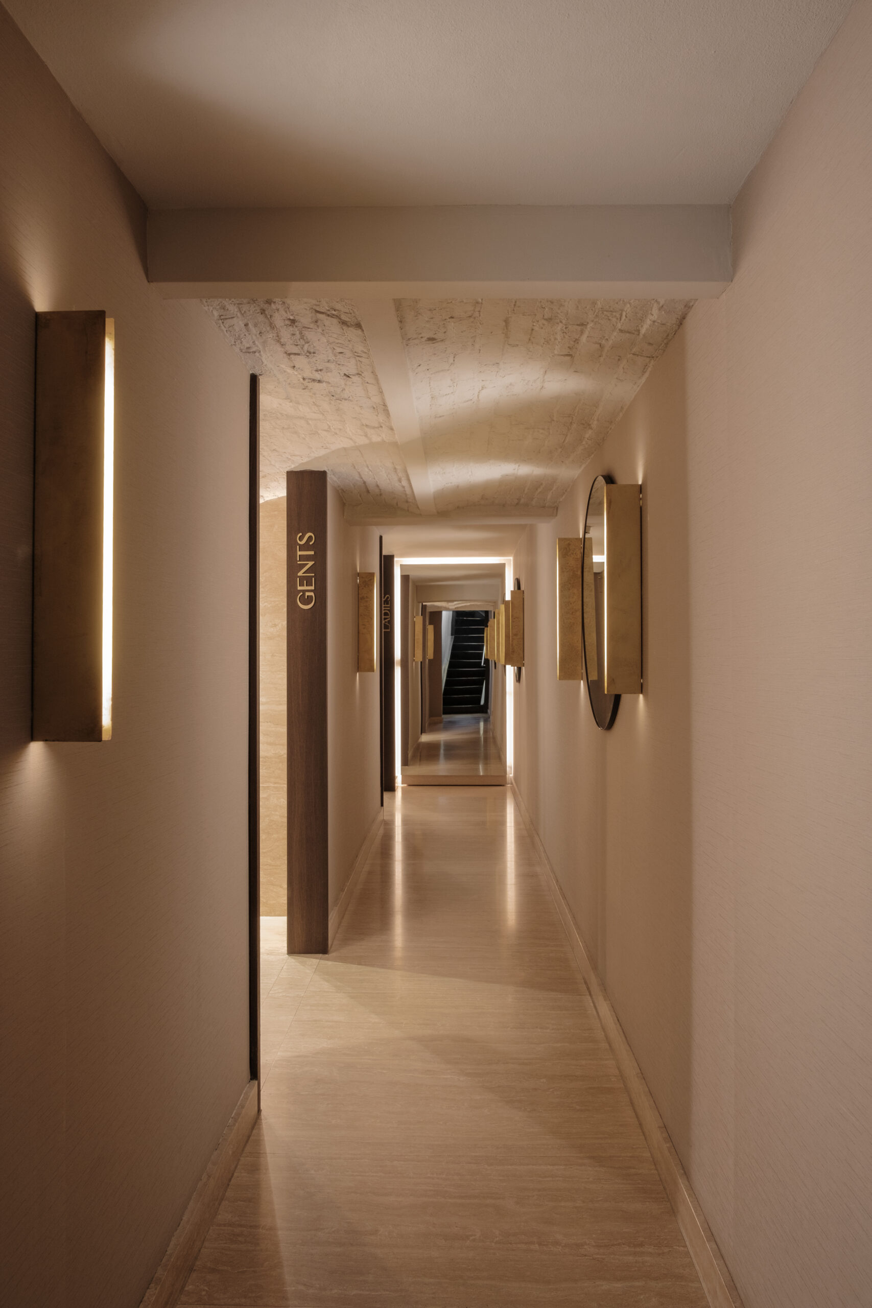 Gang naar de herentoiletten, met gewelfd plafond, wandlampen en spiegels richting een trap